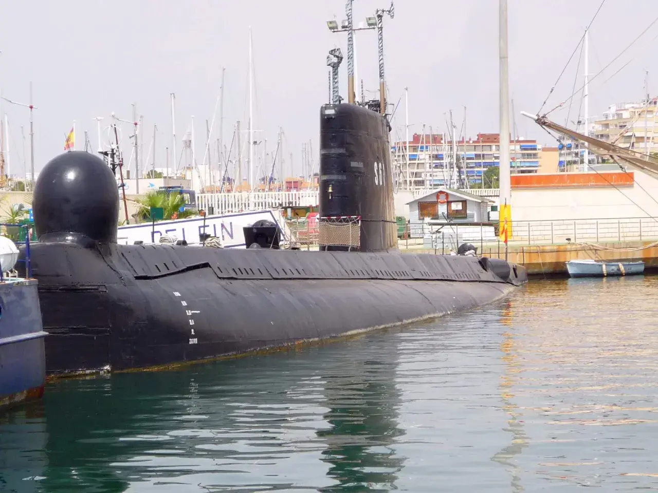 Un submarino negro, el Delfín, descansa en el puerto de Torrevieja, con mástiles de veleros y edificios al fondo.