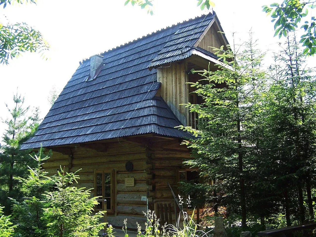 Drewniana pustelnia Brata Alberta, otoczona zielenią, z charakterystycznym gontowym dachem i kamiennym kominem.