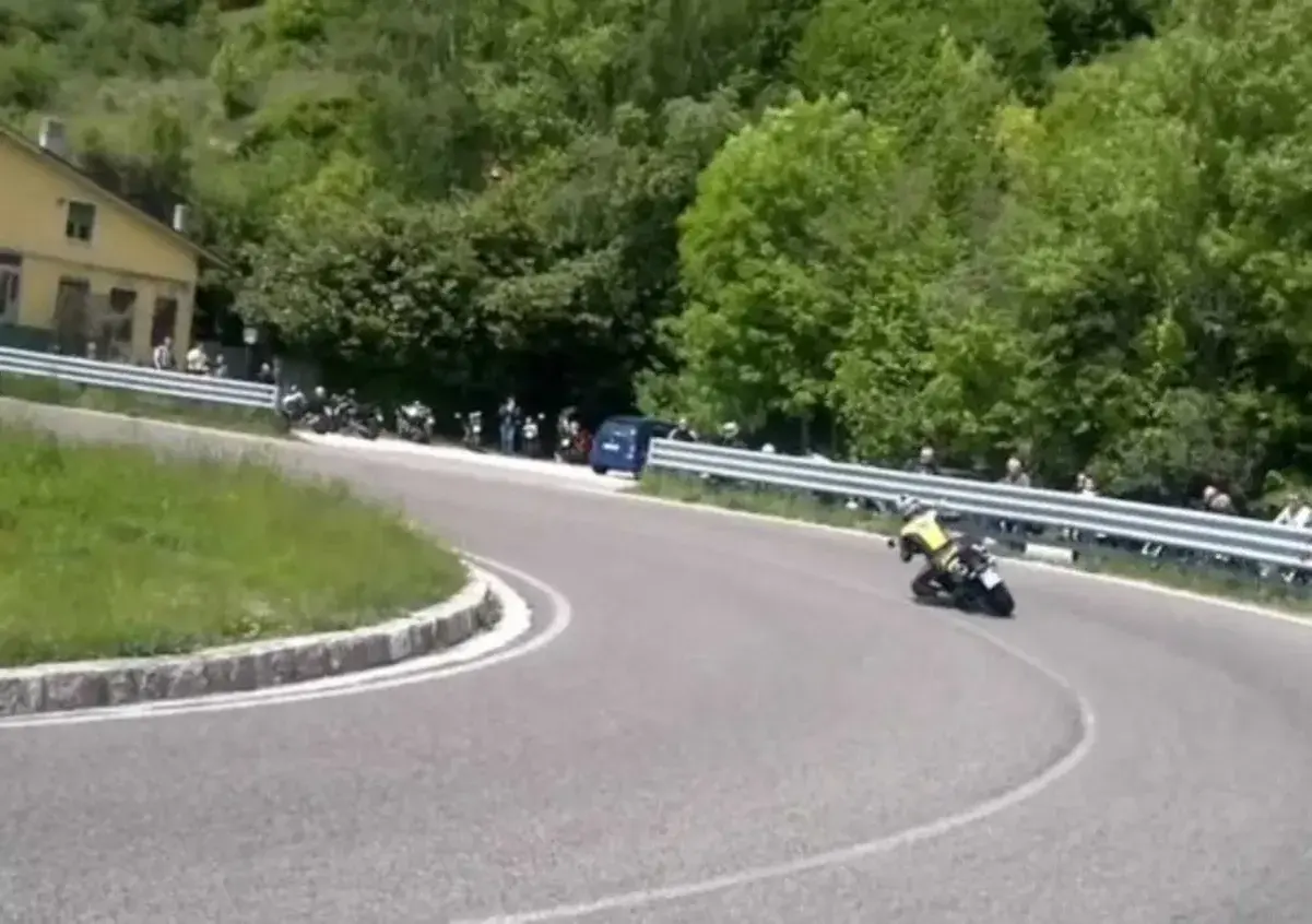 Un motociclista in giallo affronta una curva su una **strada del costo** panoramica, con spettatori e moto parcheggiate a lato.