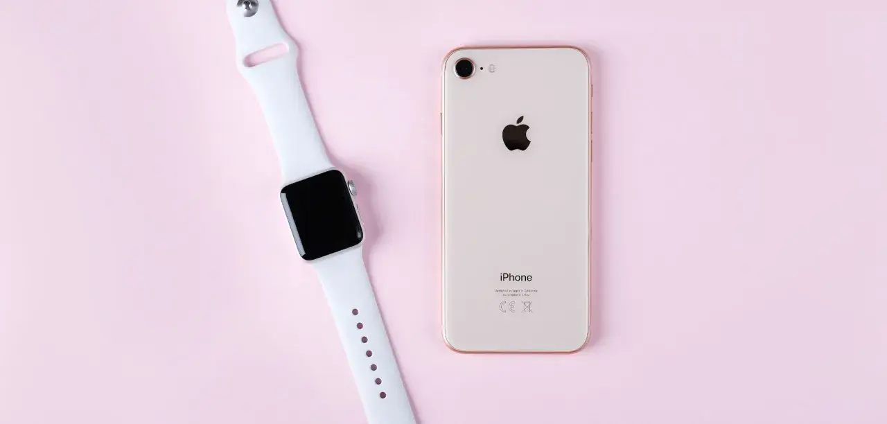 Apple Watch und iPhone liegen nebeneinander auf rosa Untergrund, bereit zum Laden.