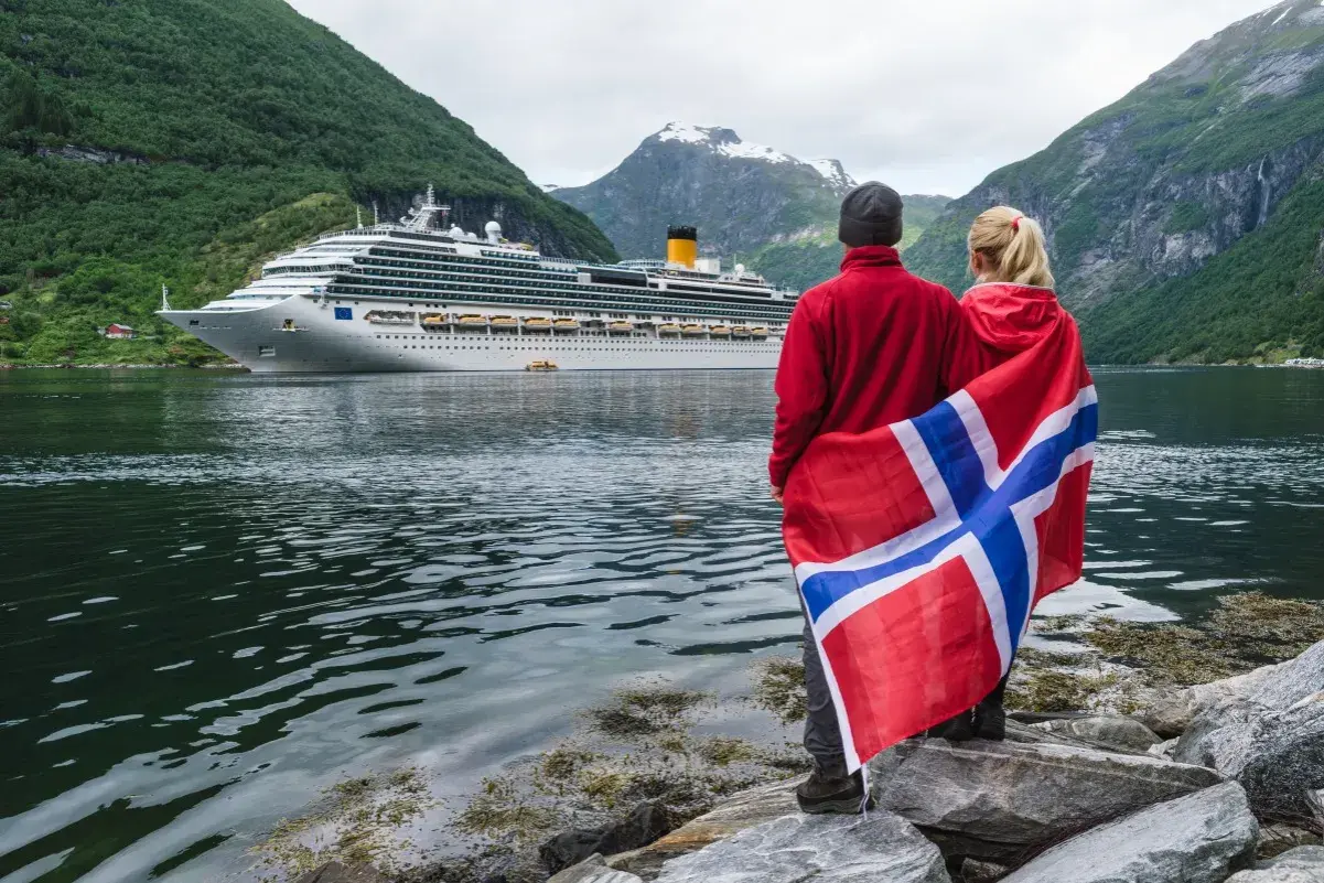 Para podróżuje po Norwegii, z flagą narodową i statkiem wycieczkowym w tle. Planujesz podróż? Pomyśl o ubezpieczenie Norwegia.