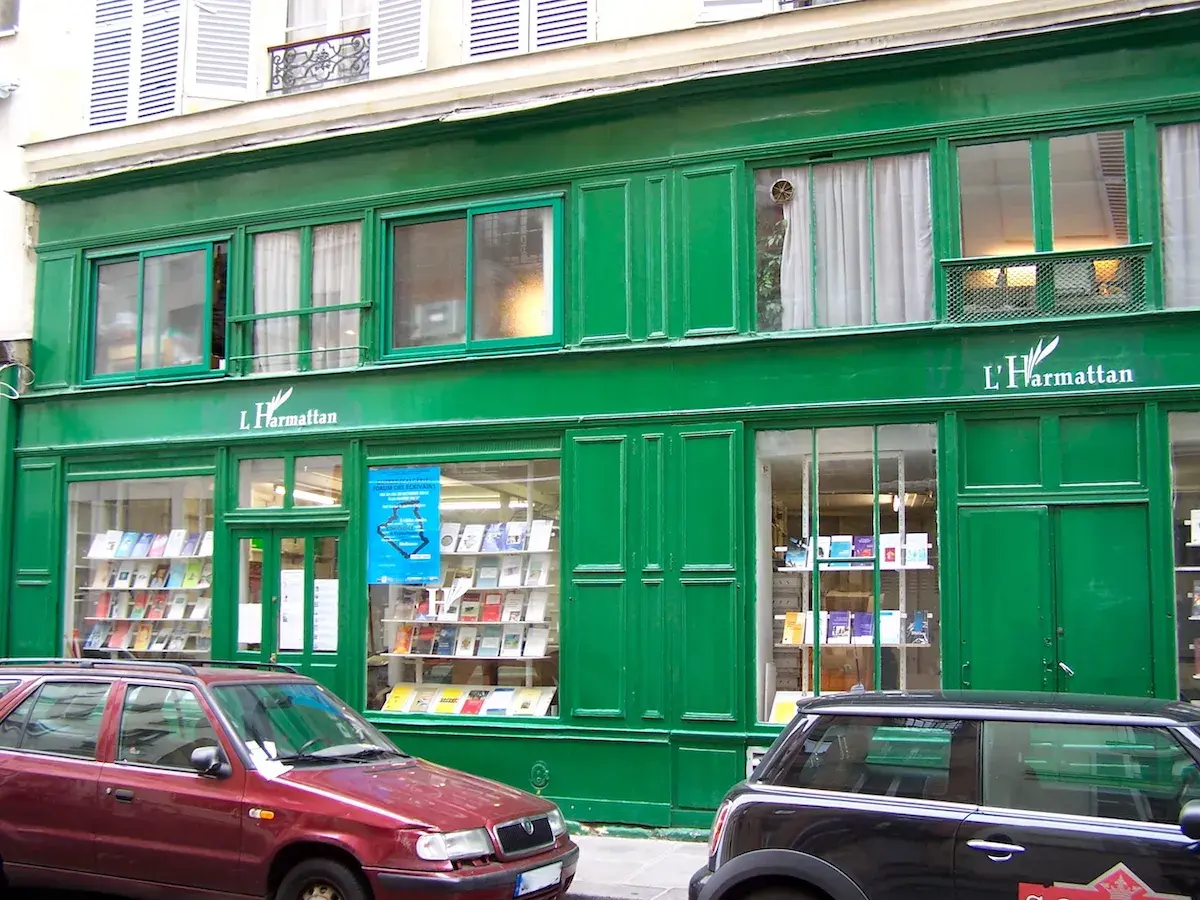 Façade verte d'une librairie L'Harmattan, vitrines remplies de livres. Une voiture rouge et une noire stationnent devant.