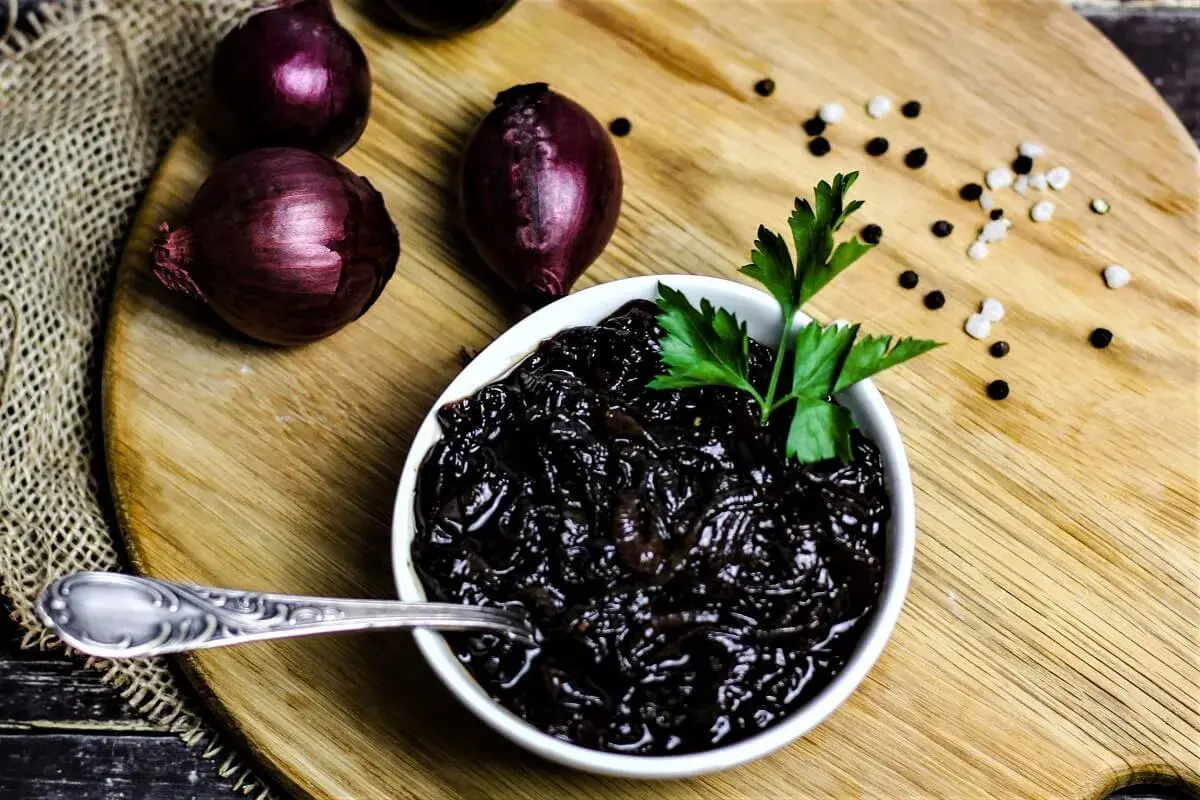Schwarze, karamellisierte balsamico zwiebeln in einer Schüssel, garniert mit Petersilie. Daneben rote Zwiebeln und Pfefferkörner.