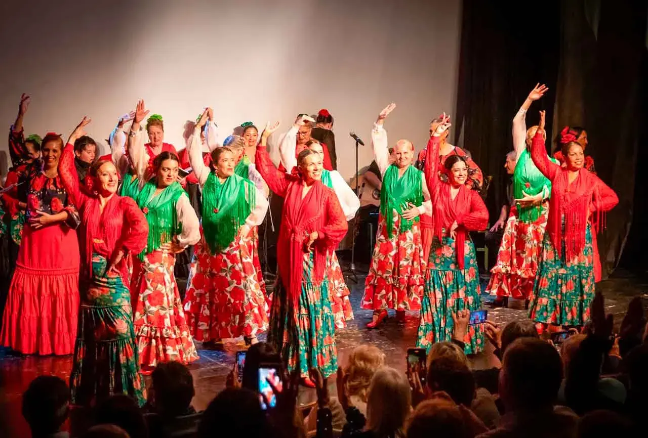 Un grupo de mujeres baila sevillanas de amor con trajes coloridos. Sus faldas amplias y mantones vibrantes llenan el escenario de alegría.