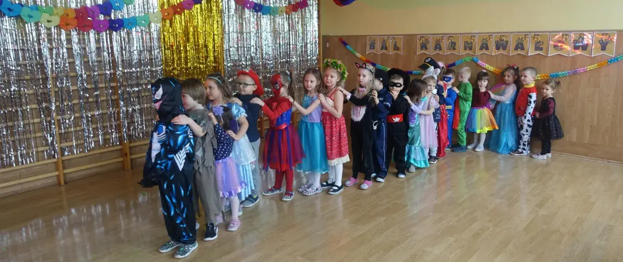 Dzieci w strojach karnawałowych stoją w kolejce na tle błyszczących zasłon i dekoracji.