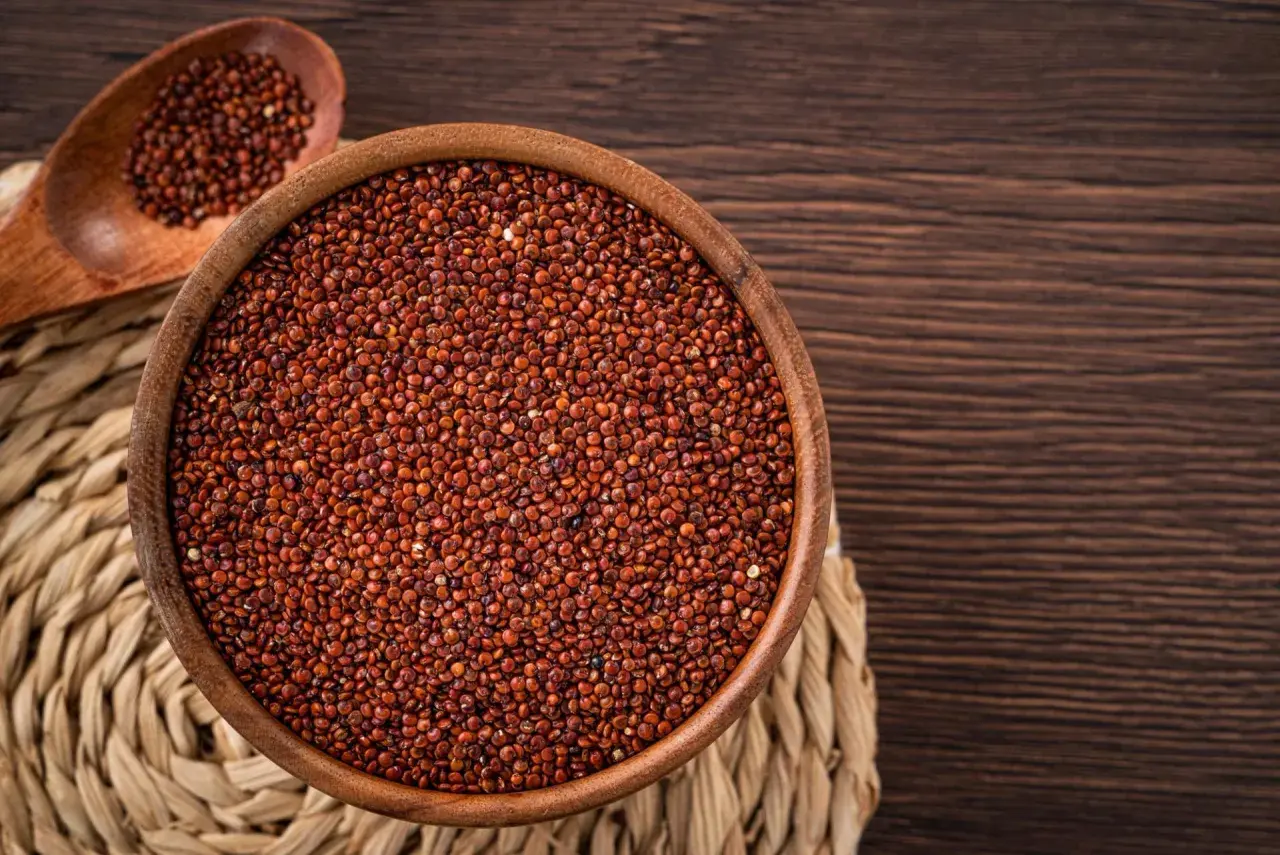 Una ciotola di legno piena di quinoa rossa, con un cucchiaio di legno accanto, su un sottobicchiere intrecciato.