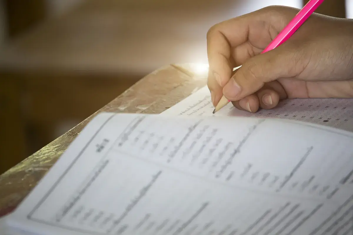 Dłoń ucznia pisząca różowym ołówkiem na arkuszu testowym z kółkami do zaznaczenia odpowiedzi.