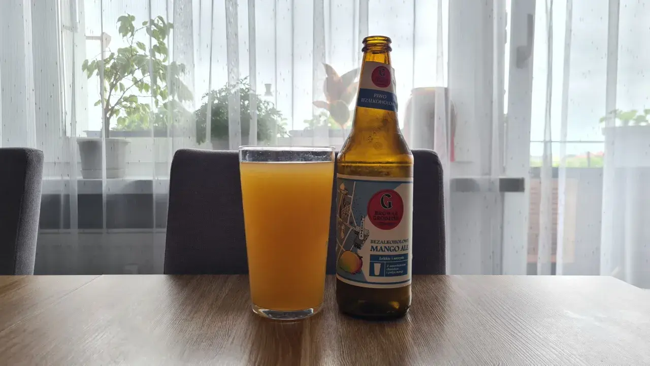 Szklanka z pomarańczowym napojem i butelka piwa bezalkoholowego mango ale na stole.