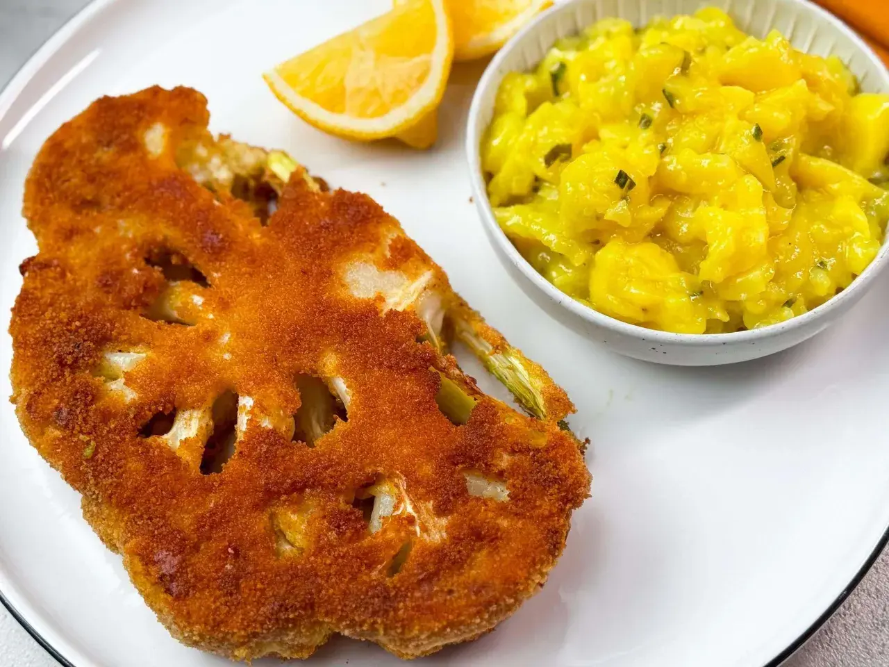 Knuspriges Blumenkohl-Schnitzel mit Kartoffelsalat und Zitronenspalten auf einem weißen Teller.