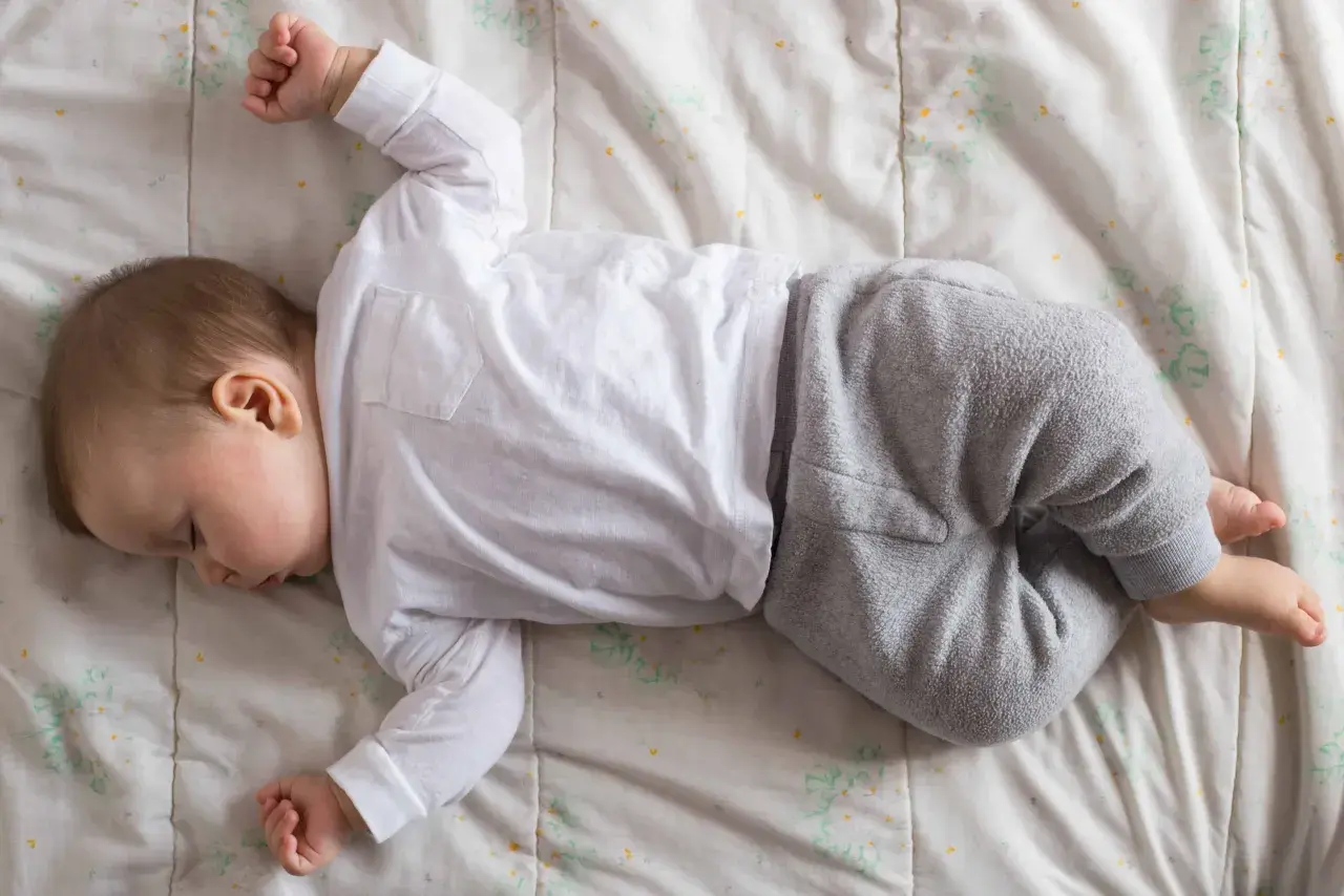 Un neonato non dorme, ma riposa sul fianco, vestito di bianco e grigio, su un lenzuolo con fiori stilizzati.
