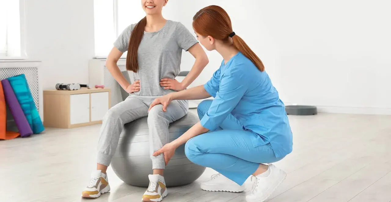 Une kinésithérapeute aide une patiente assise sur un ballon de gym à retrouver de la force dans les jambes grâce à des exercices ciblés.