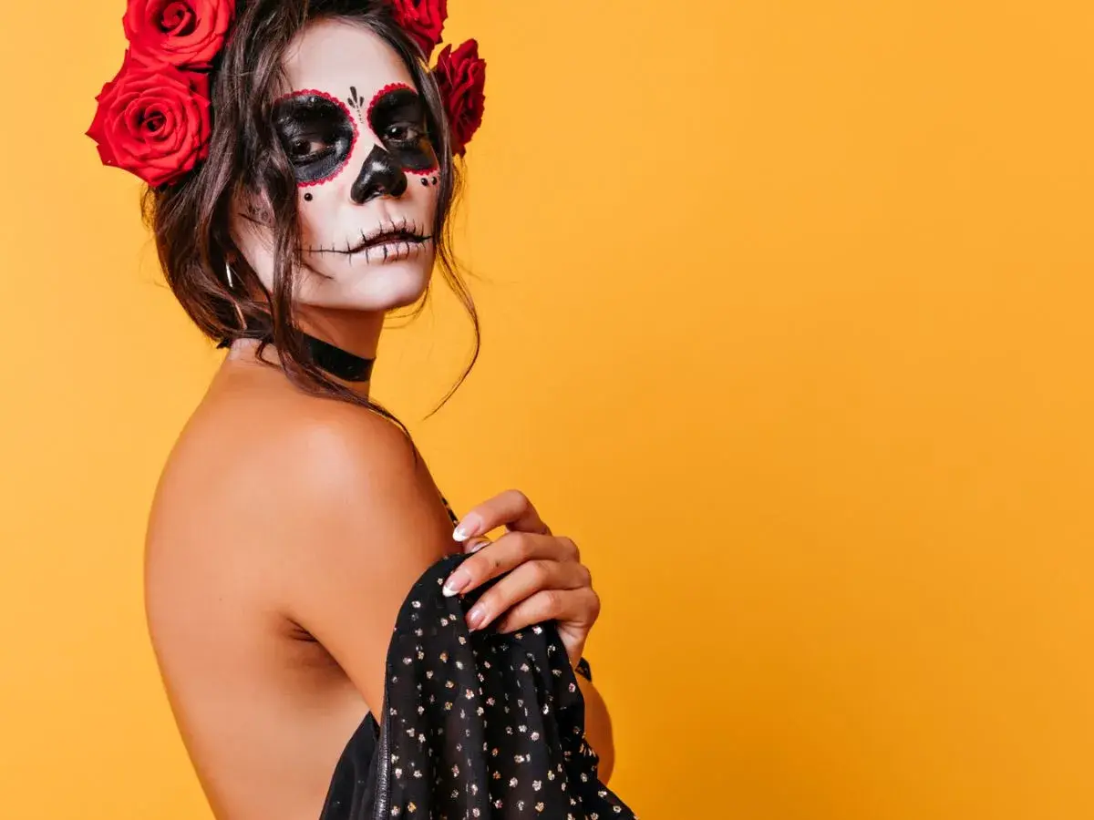 Kobieta z makijażem sugar skull i czerwonymi różami we włosach na pomarańczowym tle.