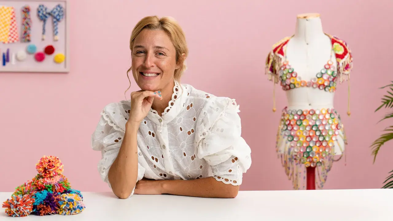 Mujer sonriente junto a un maniquí con un traje colorido de upcycling moda. Montón de pompones de colores a la izquierda.