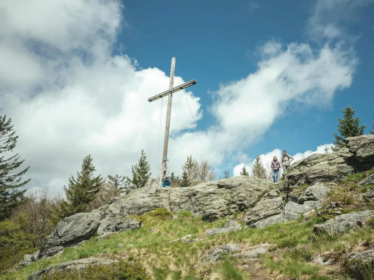 Wanderer auf einem felsigen Gipfel im Bayerischen Wald unter einem Holzkreuz. Ein typischer 8-Tausender-Ausblick.