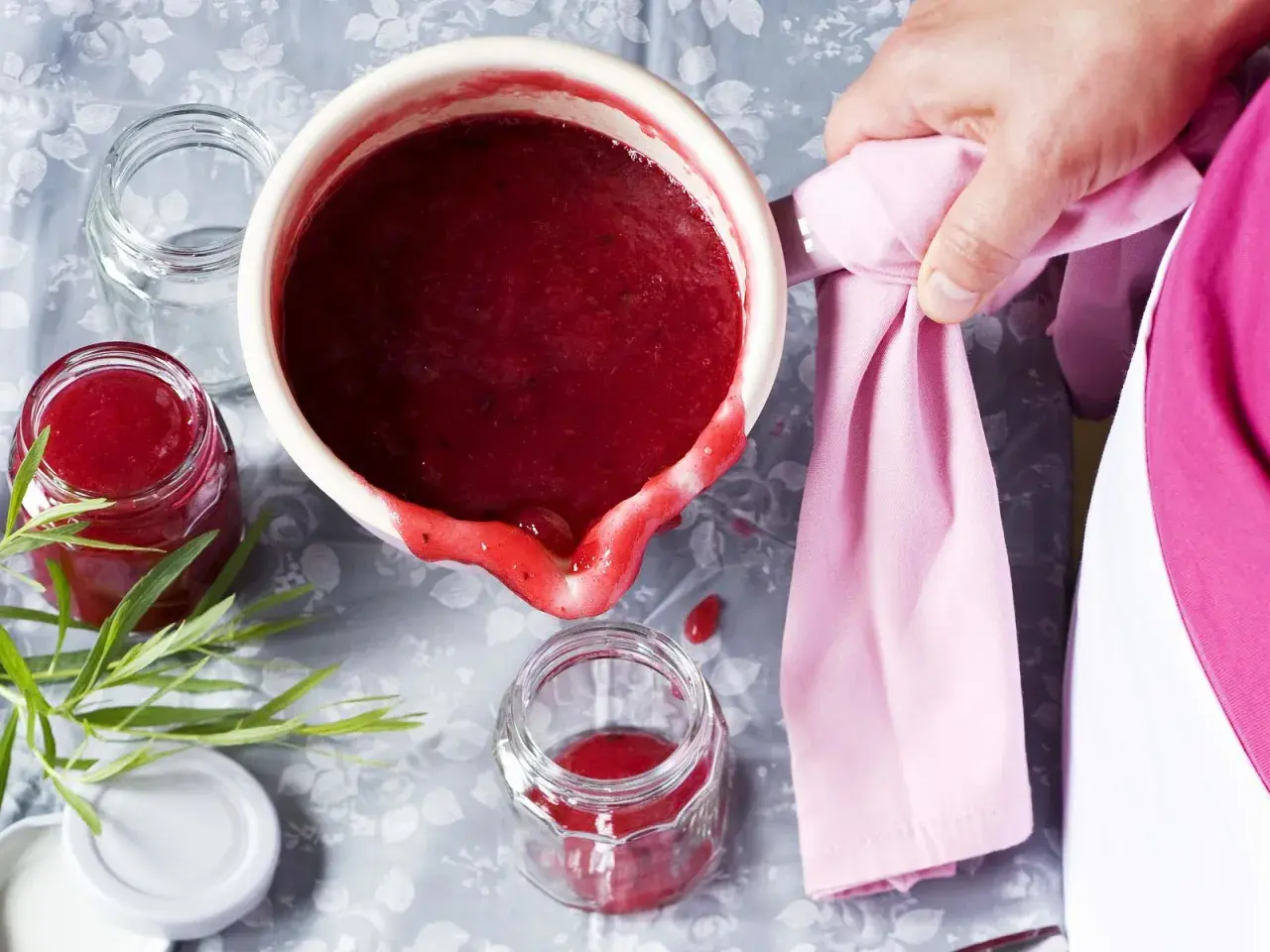 Frisch gekochte, einfache Johannisbeermarmelade wird in ein Glas gefüllt. Ein Hauch von Grün und ein rosa Tuch vervollständigen das Bild.