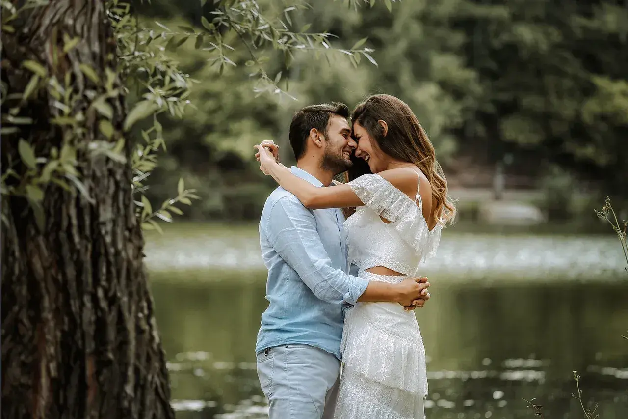 Para młoda, w białej sukni i błękitnej koszuli, przytula się czule nad jeziorem. Ich uśmiechy i spojrzenia sugerują szczęśliwą noc poślubną.