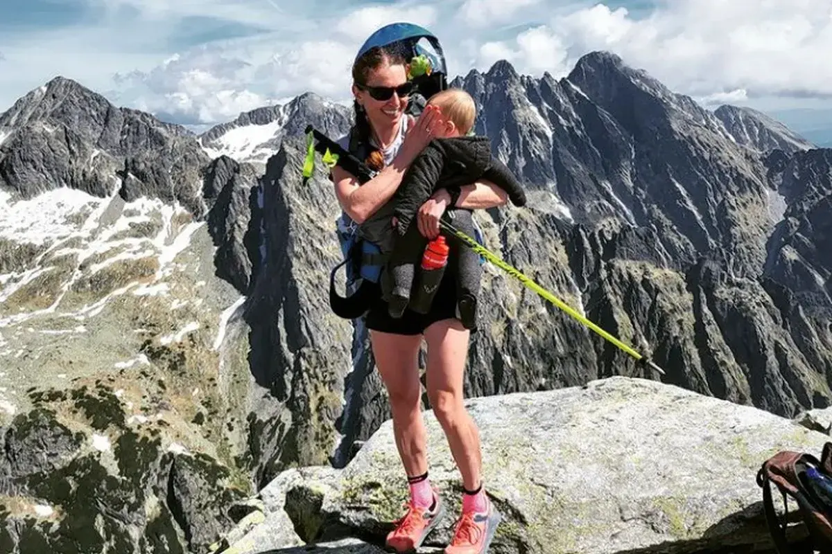 Justyna Kowalczyk z dzieckiem na szczycie górskim, w tle majestatyczne, skaliste pasmo.
