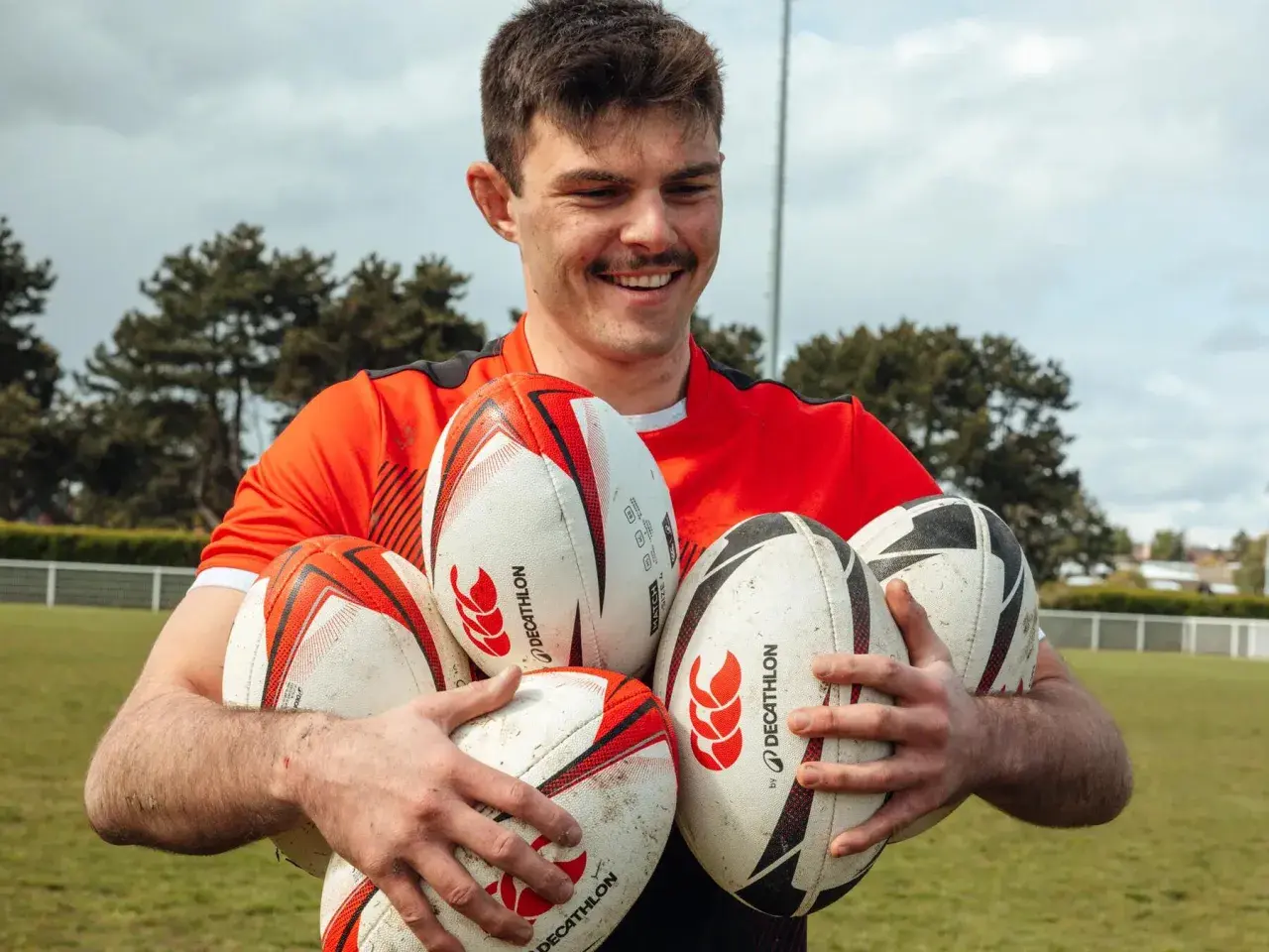 Uśmiechnięty mężczyzna w czerwonej koszulce trzyma kilka piłek do rugby i futbolu amerykańskiego.