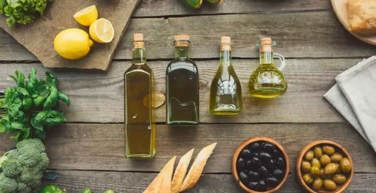 Botellas de aceite refinado de oliva junto a limones, hierbas frescas, pan y aceitunas sobre una mesa de madera.