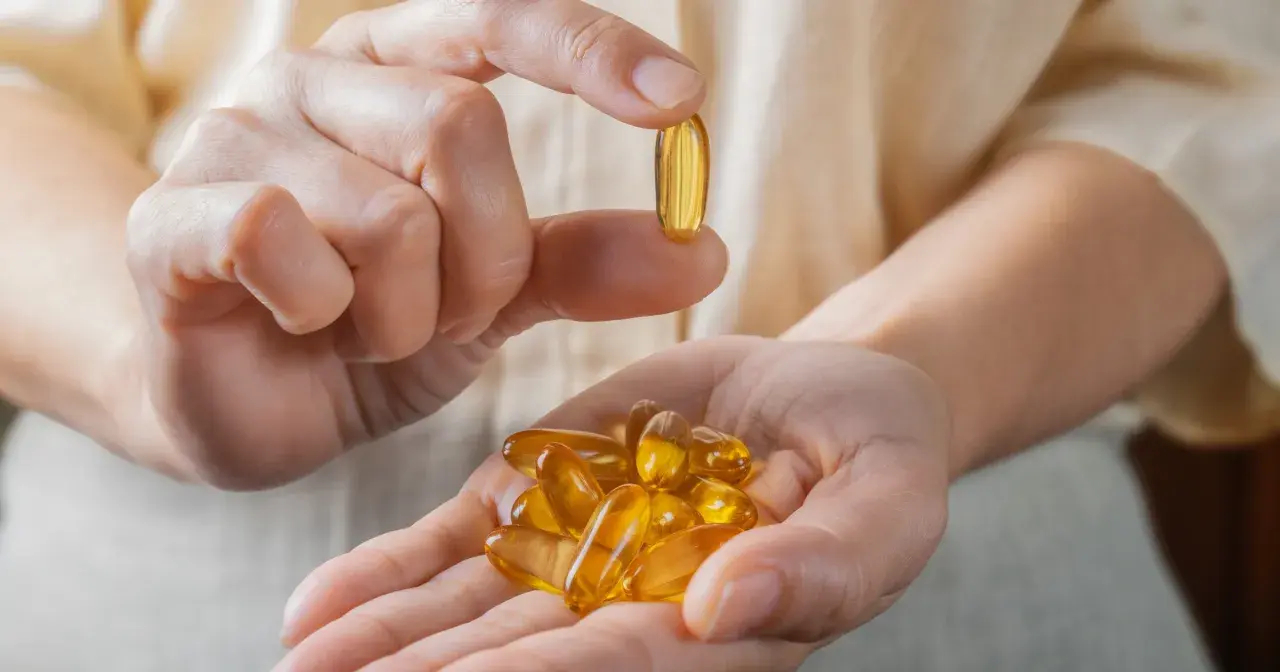 Dłoń trzyma garść kapsułek z olejem rybim, bogatym w kwasy omega 3. Jedna kapsułka jest podniesiona między palcami.
