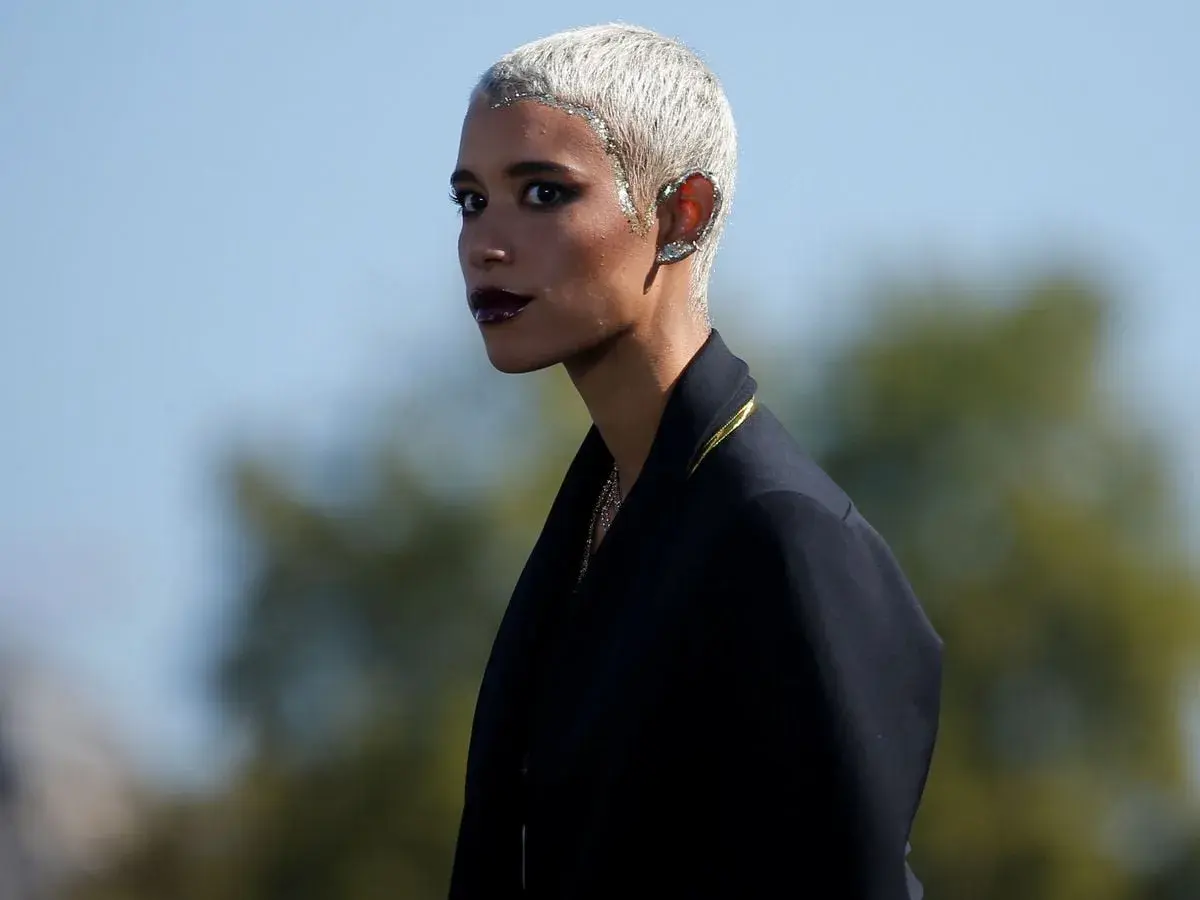 Mujer con **pelo blanco** corto y brillante, maquillaje oscuro y un pendiente llamativo en la oreja.