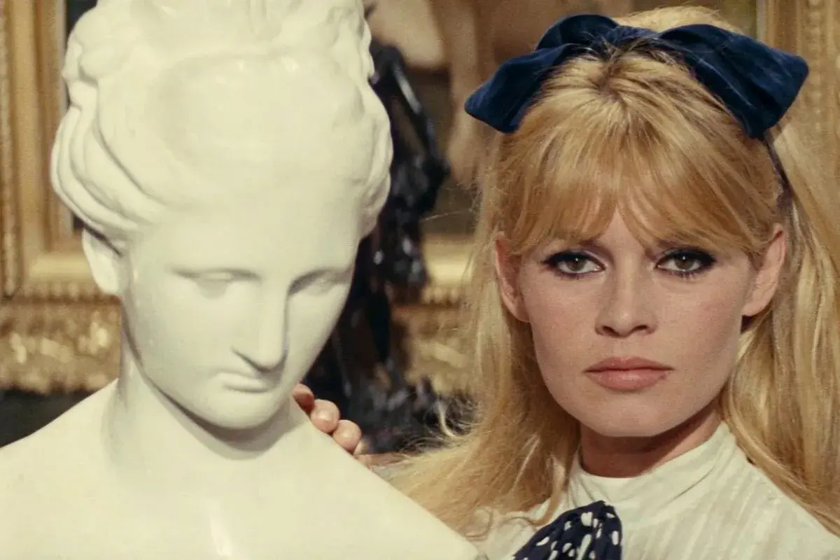 Brigitte Bardot z niebieską kokardką we włosach, obok antycznej rzeźby. Ikona lat 60 muzyka i stylu.