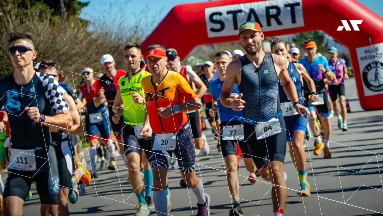 Zawodnicy biegną pod czerwoną bramką startową. Wszyscy przestrzegają zasad triathlonu, skupieni na rywalizacji.