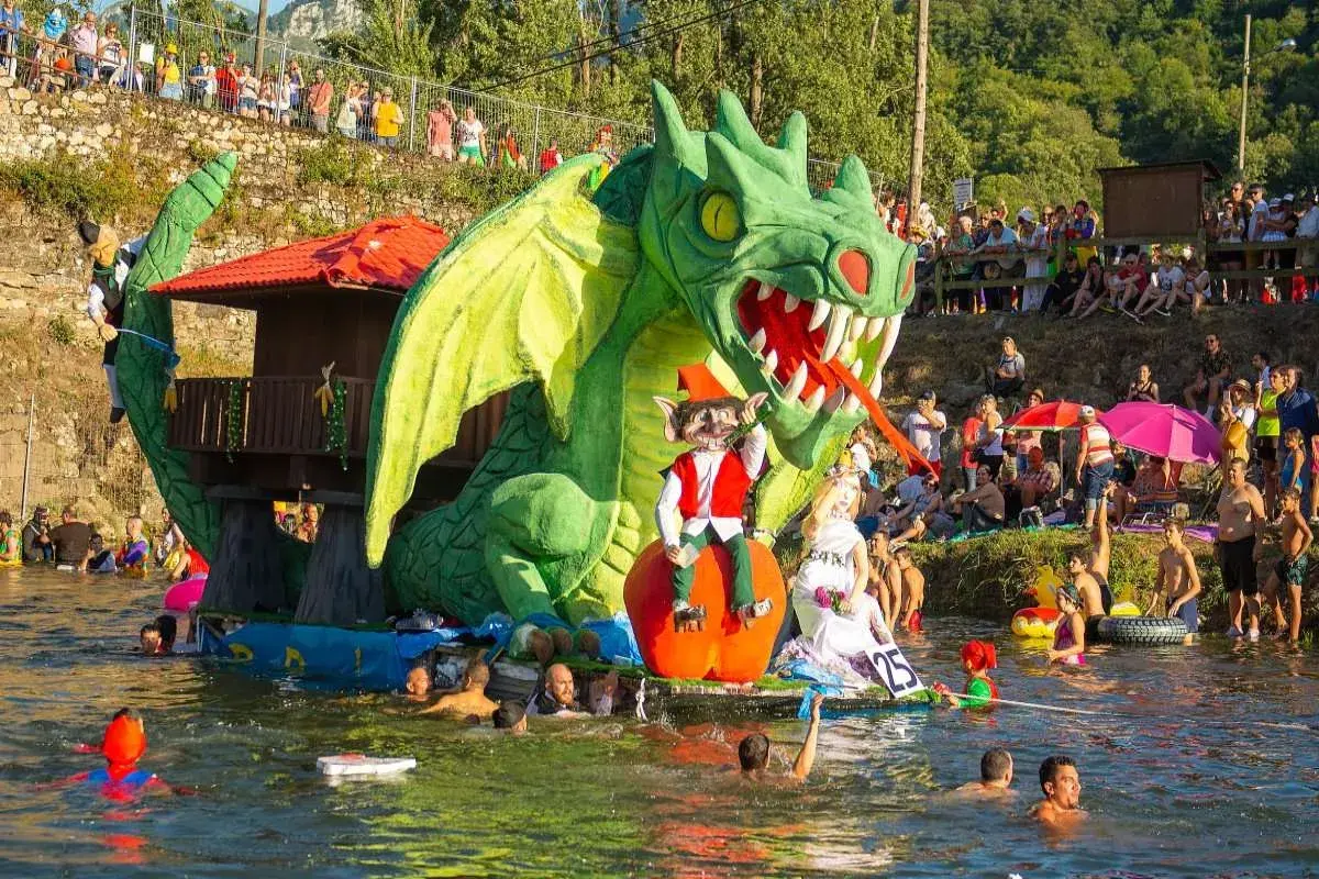 Dragón verde gigante en un desfile acuático, parte de las fiestas de prao Asturias. Gente en el agua y espectadores en la orilla.