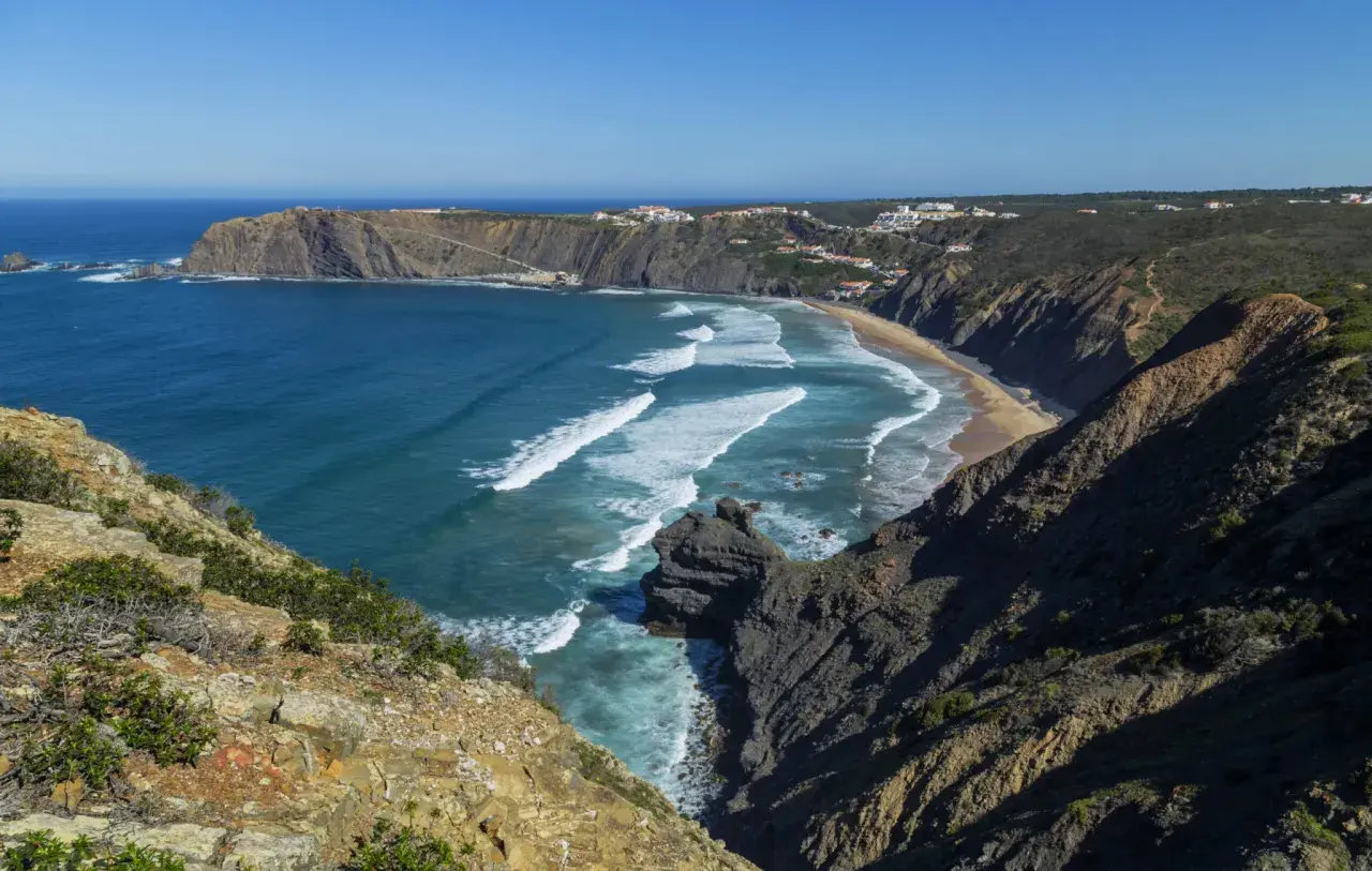 Malownicze wybrzeże Portugalii z klifami, piaszczystą plażą i turkusowym oceanem. Białe fale rozbijają się o brzeg, a w oddali widać białe domki.