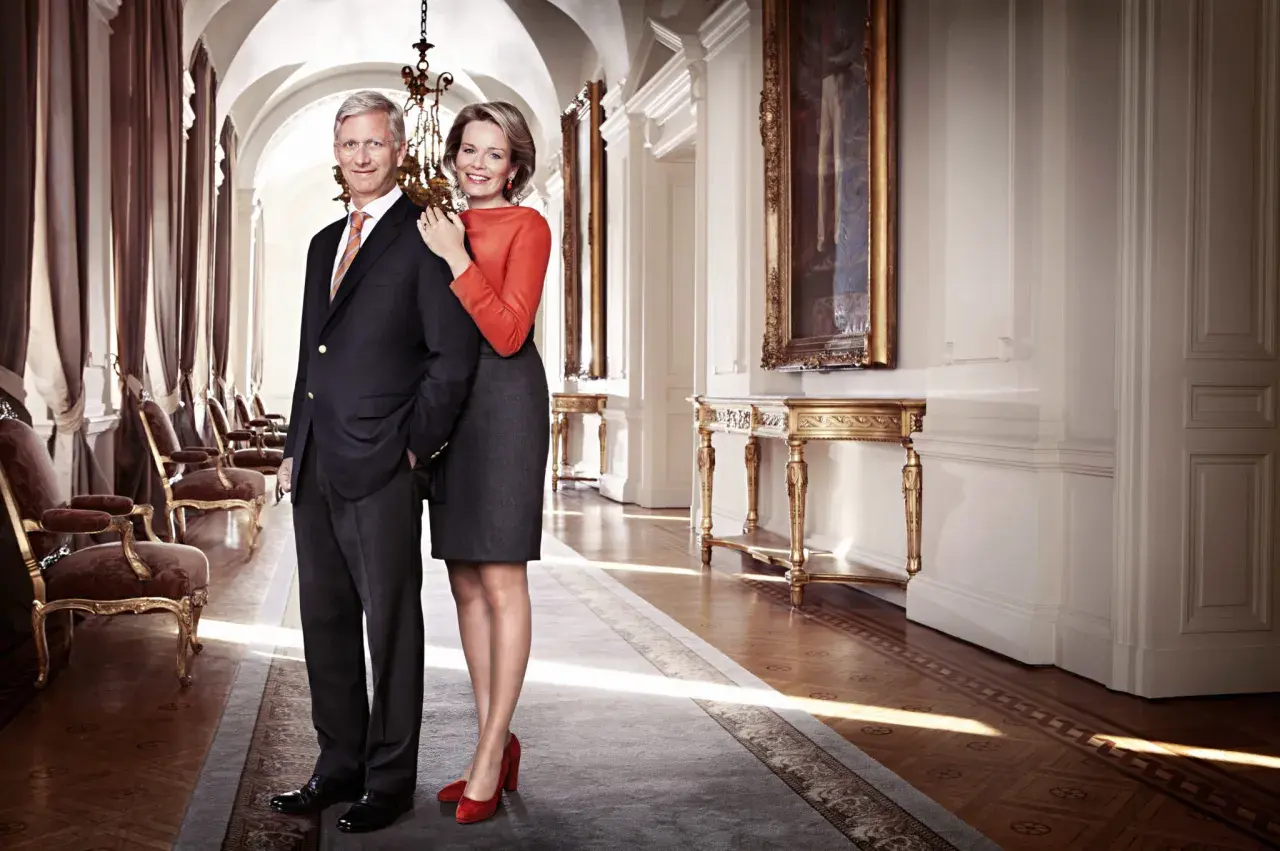 Królowa Belgii, uśmiechnięta, stoi obok króla w eleganckim korytarzu pałacowym.