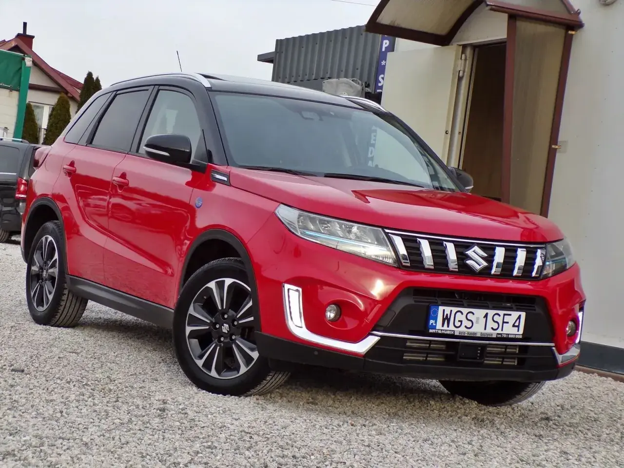 Czerwony Suzuki Vitara Hybrid z czarnym dachem, zaparkowany na żwirowej nawierzchni.
