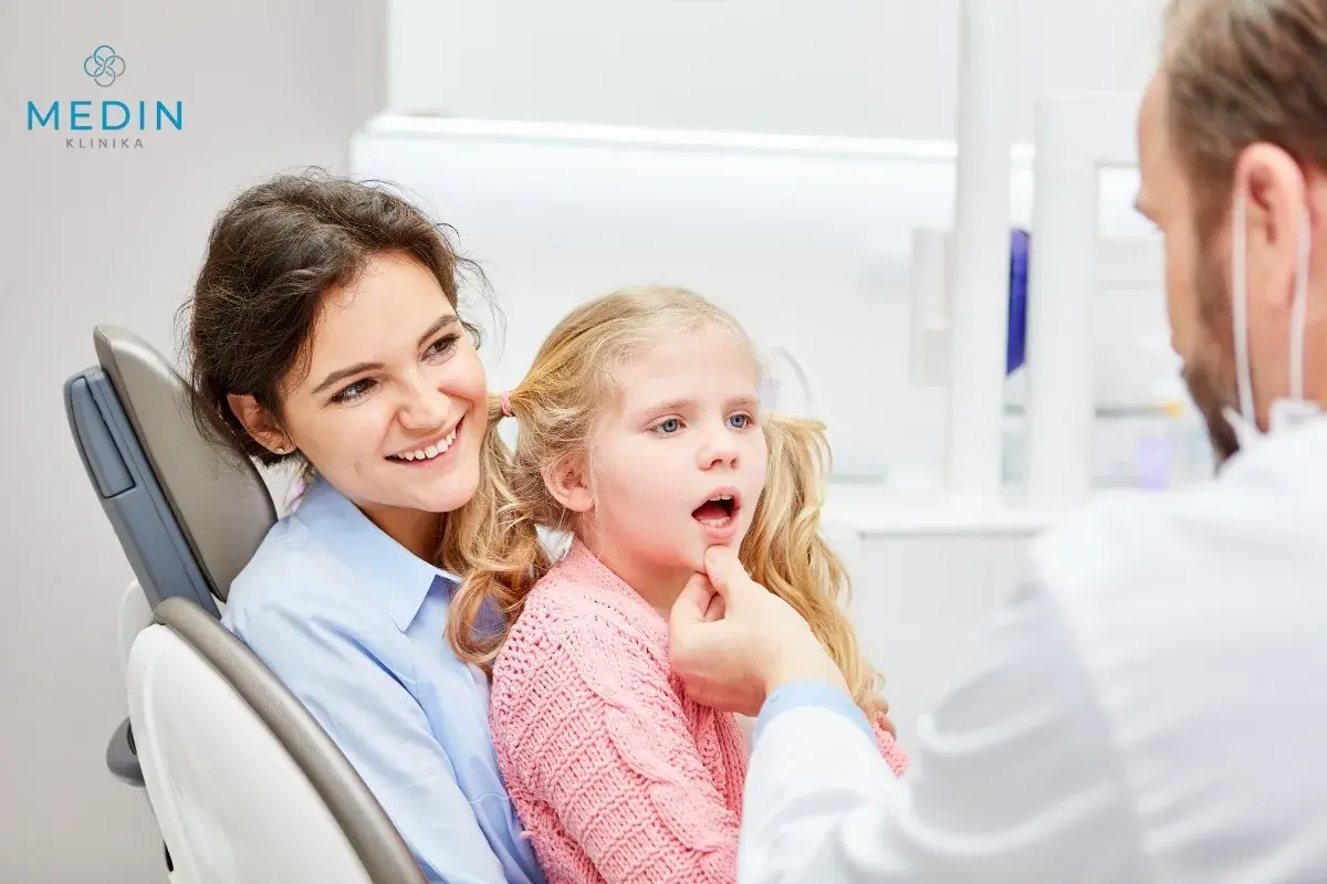 Uśmiechnięta mama towarzyszy córce podczas zabiegu lakowania zębów u dzieci. Dentysta bada jamę ustną dziewczynki.