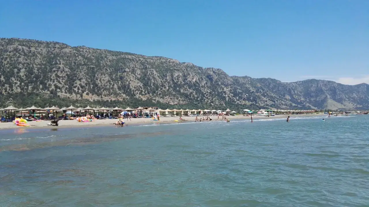 Słoneczny dzień na plaży Velipoja. Ludzie odpoczywają pod parasolami, kąpią się w morzu, a w tle wznoszą się górzyste tereny.