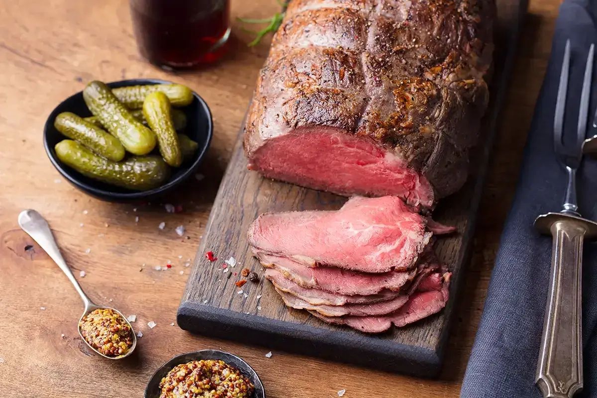 Un rôti de bœuf tranché, servi avec des cornichons, de la moutarde et des couverts sur une planche en bois.