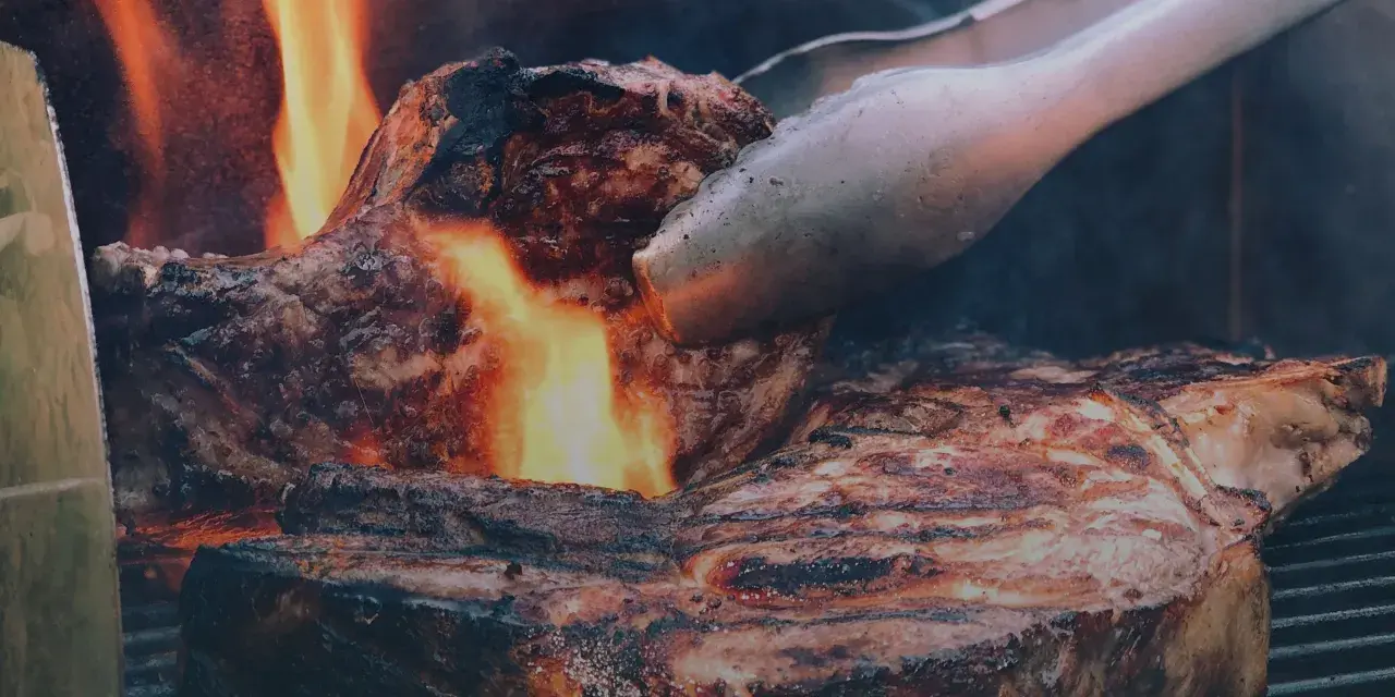 En saftig entrecote grillas över öppen eld, perfekt innertemp. Tången vänder köttet för en jämn stekyta.