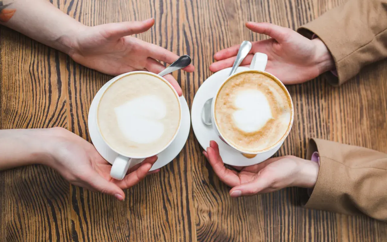 Zwei Cappuccinos mit Herz-Latte-Art, die Hände reichen sie sich über einen Holztisch. Wie viel Milch ist im perfekten Cappuccino?
