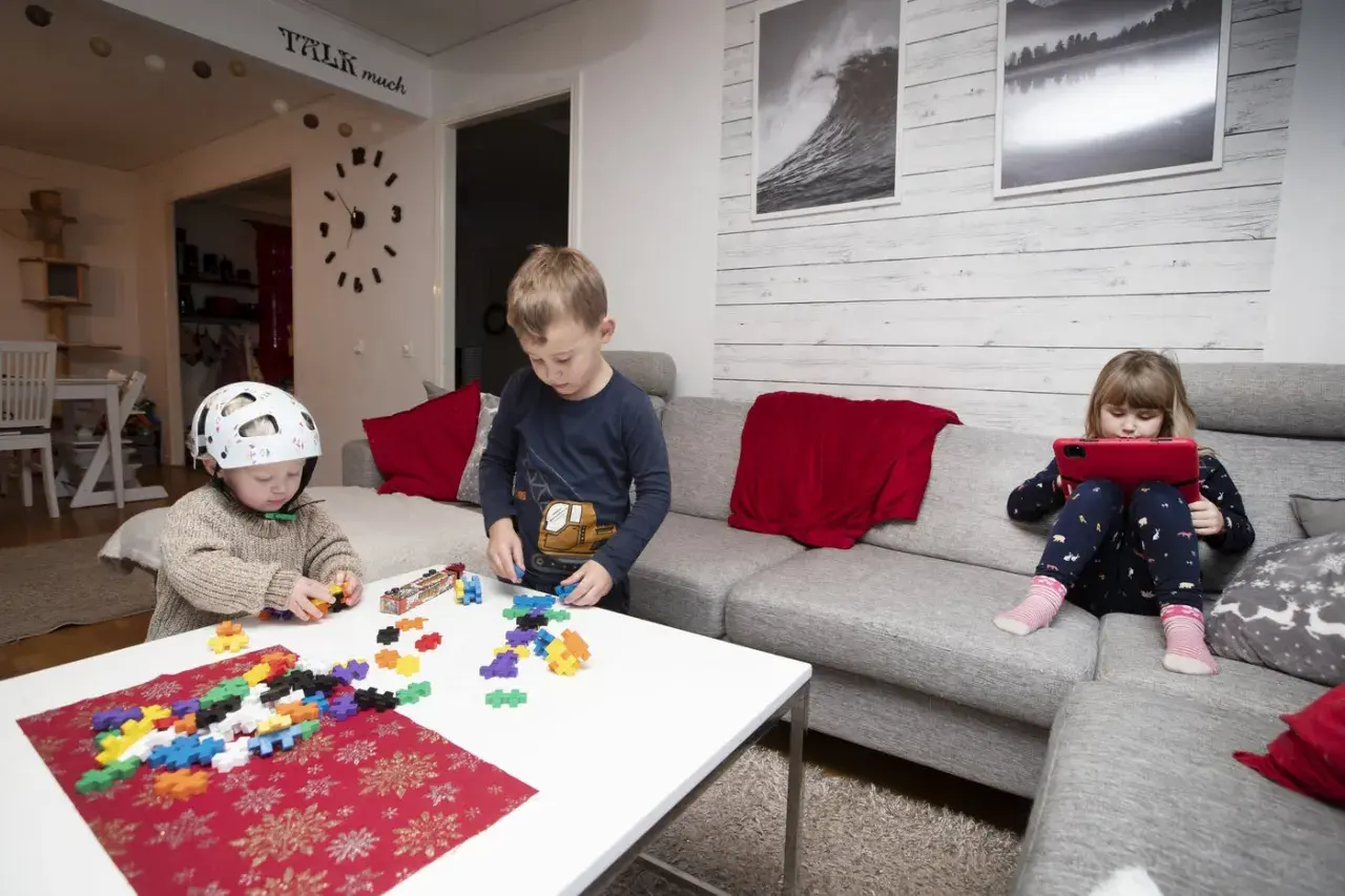 Tre barn leker med byggklossar och en surfplatta i ett vardagsrum. En pojke med hjälm bygger, en annan pojke leker med klossar, och en flicka tittar på en surfplatta. Leo Carmona barnen har kul.