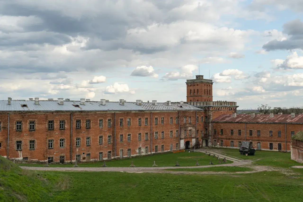 Dawny fort wojskowy w okolicach Warszawy, z ceglanymi budynkami i wieżą.