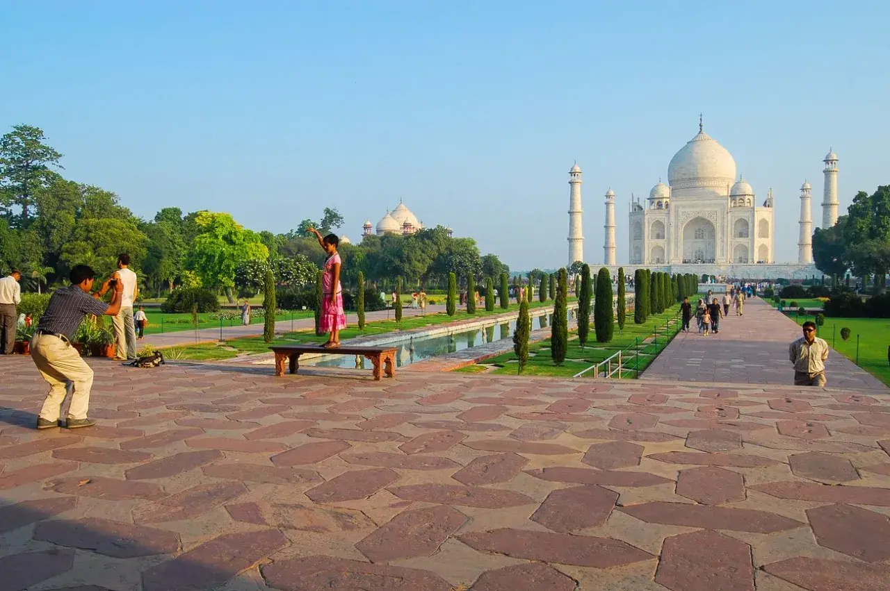 Ein Mann fotografiert ein Mädchen, das auf einer Bank vor dem Taj Mahal steht. Die Anlage des Taj Mahal Land ist mit Bäumen und einem Wasserbecken geschmückt.