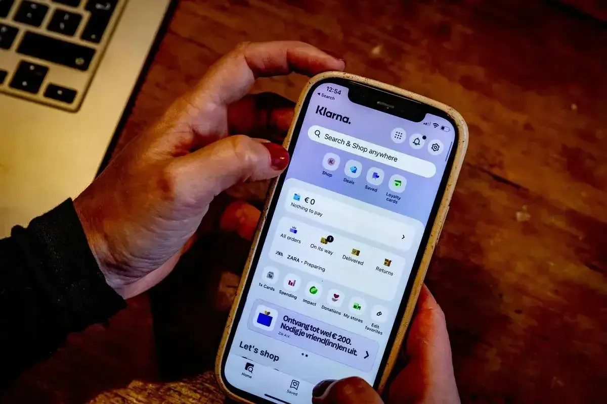 Dłoń trzyma smartfon z aplikacją Klarna. Czy Klarna jest bezpieczna? Aplikacja oferuje zakupy, płatności i zarządzanie zamówieniami.