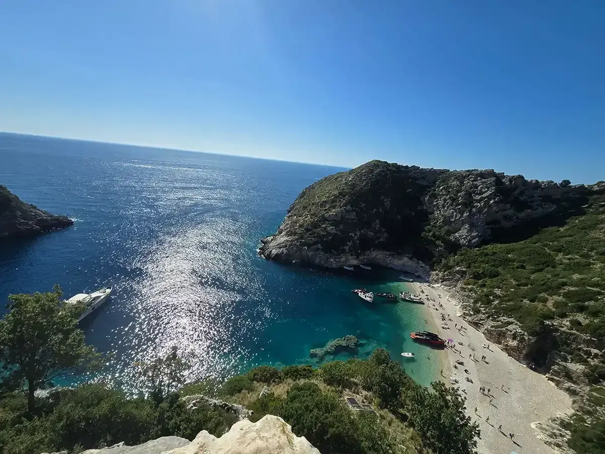 Słoneczna zatoka grama z turkusową wodą, kamienistą plażą pełną ludzi i łodziami przycumowanymi do brzegu.
