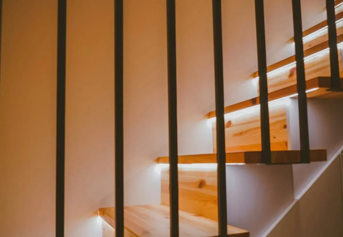 Drewniane stopnie schodów z subtelnym oświetleniem LED w ścianie. Czarne, pionowe balustrady tworzą nowoczesny kontrast.