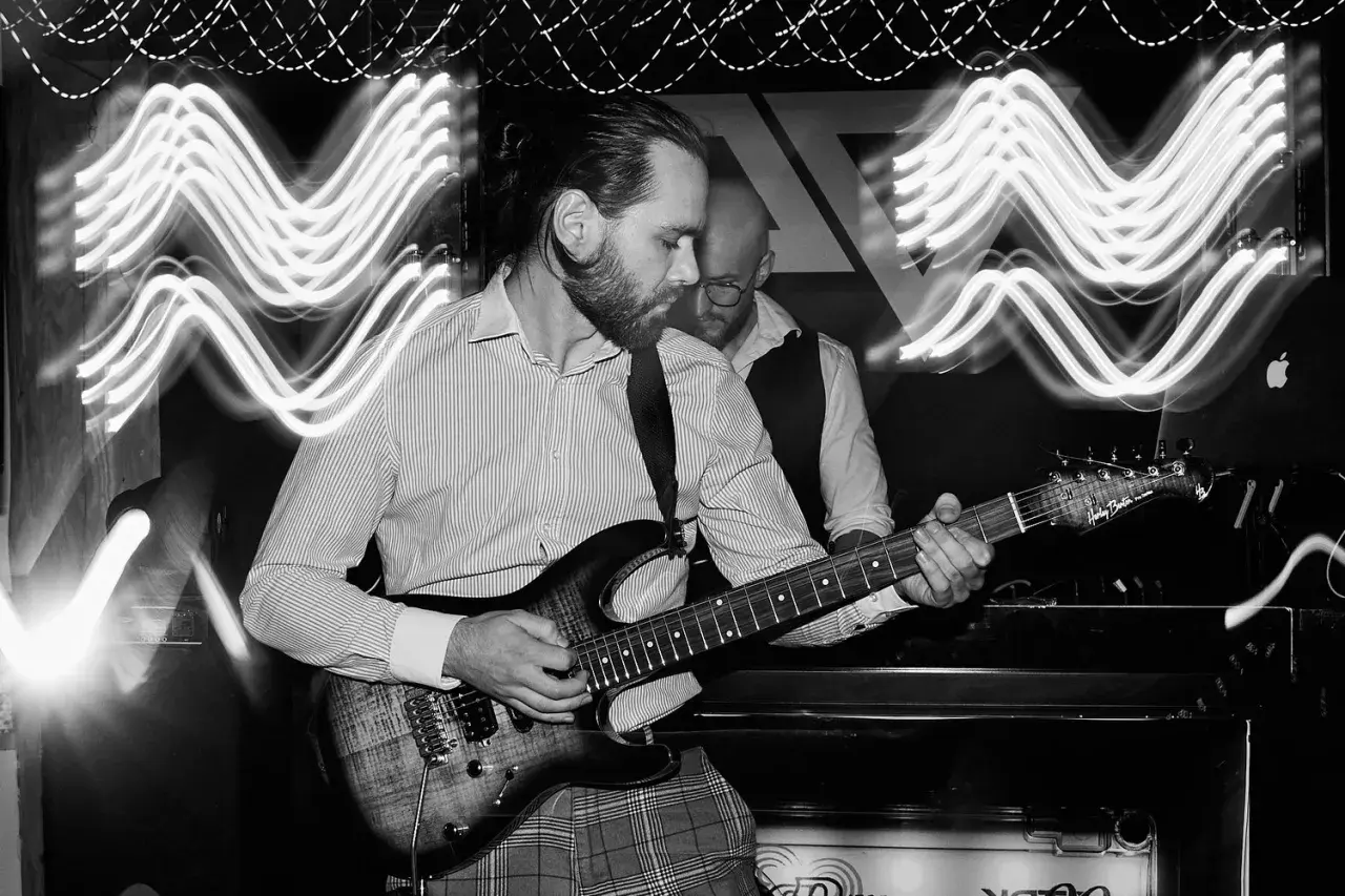Muzyk gra na gitarze elektrycznej, a za nim widać drugiego członka zespołu. Efekt falujących świateł sugeruje kompensację ekspozycji.