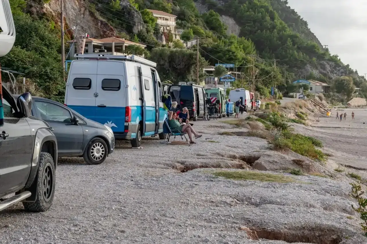 Une rangée de camping-cars en Grèce, certains garés sur un chemin de gravier près d'une plage. Des gens se détendent à l'extérieur.