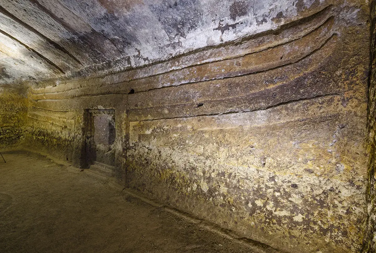 Interno di una domus de janas s'incantu, con pareti scavate e gradini che conducono a un'apertura.