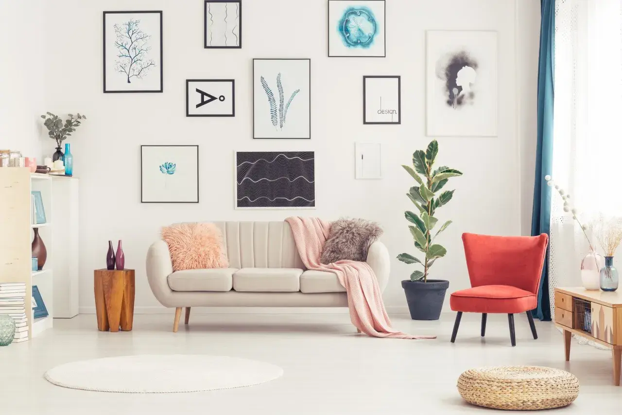 Sofa w kolorze ecru z różowymi poduszkami i pledem, obok czerwony fotel i duża roślina.