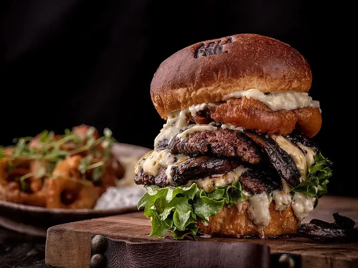 Un généreux burger maison avec plusieurs steaks, fromage fondu et laitue fraîche, servi sur une planche en bois.
