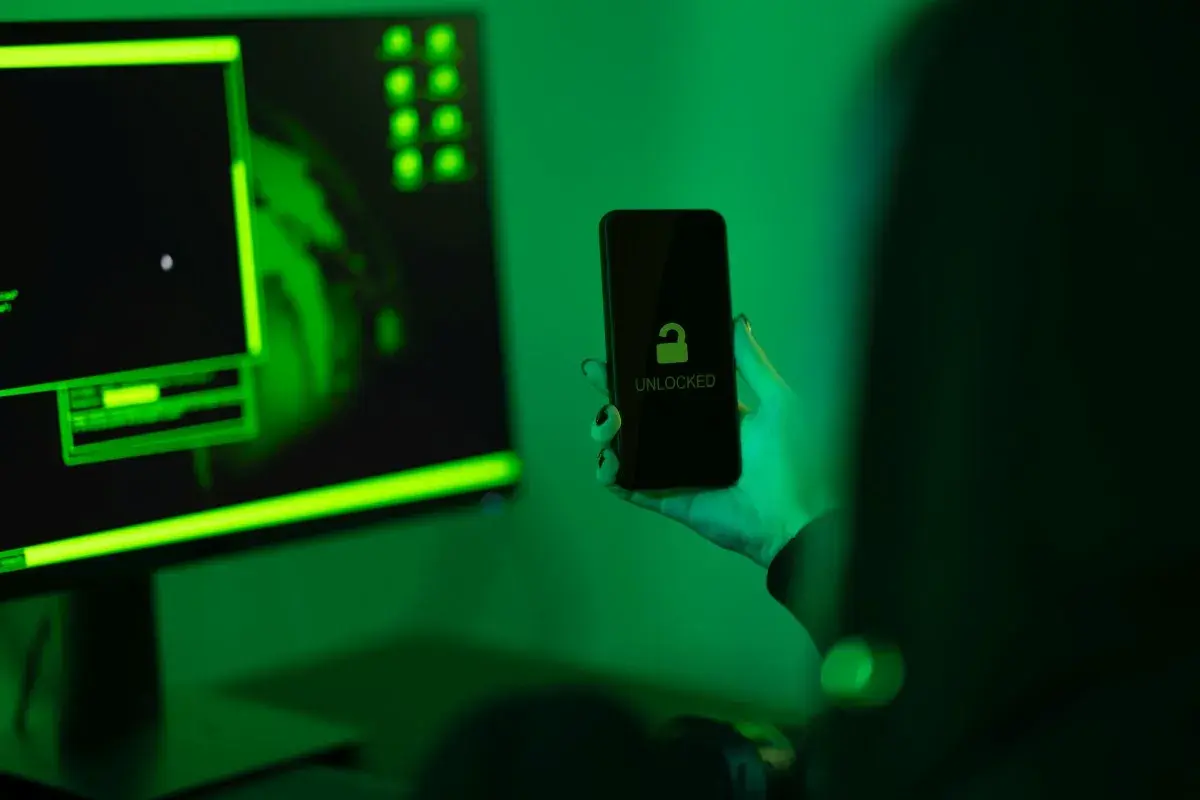 Ręka trzyma odblokowany telefon na tle monitora z zielonym kodem. Jak zrobić, by policja nie namierzyła telefonu?
