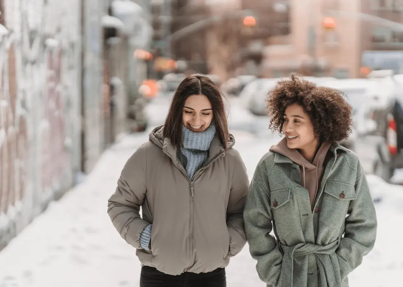 Deux amies discutent en marchant dans la neige. Une idée outfit hiver parfaite : doudoune beige et manteau vert ceinturé.