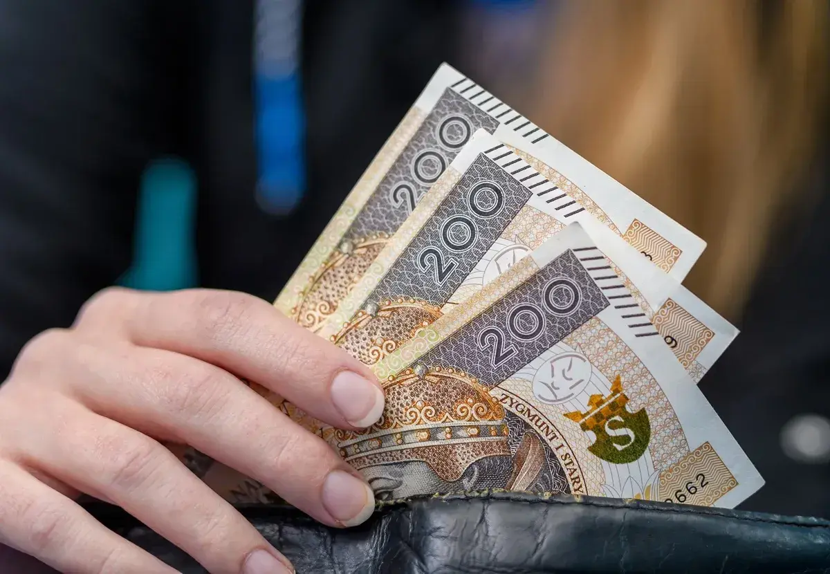 Dłoń trzyma banknoty 200 zł. Może to symbolizować wsparcie finansowe, np. stały zasiłek dla bezrobotnych po 50 roku życia.